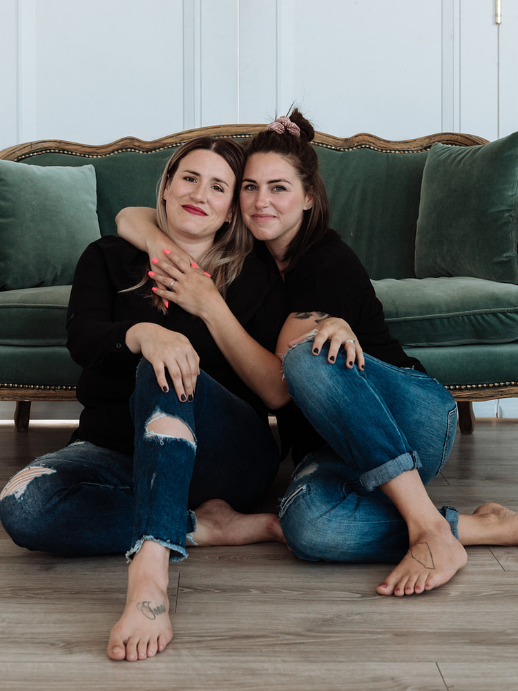 Nikki and Jenna sitting on the floor in front of a green sofa, arms wrapped around each other smiling